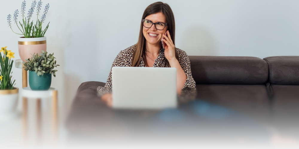 A woman on her computer making a telephone call on her sofa.